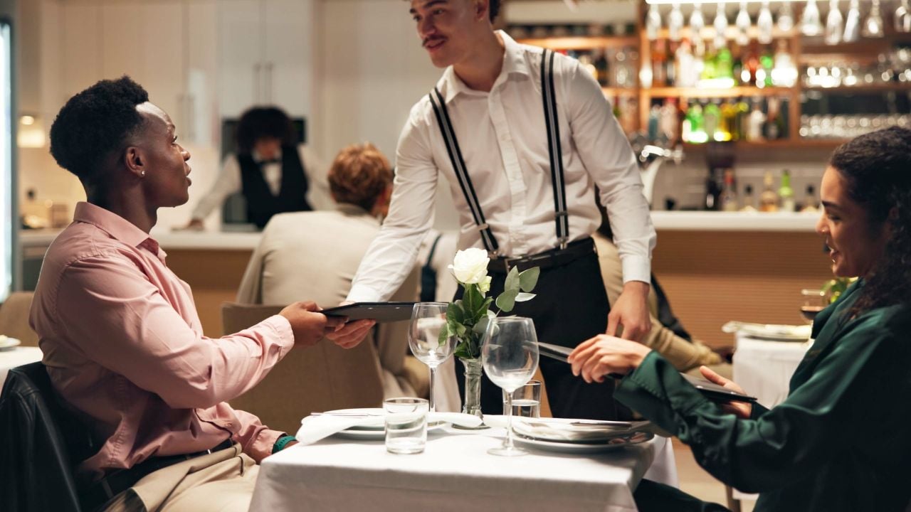 Waiter, couple and order at restaurant with menu, decision and discussion for wine list, food or service. People, server and talk for customer experience, thanks and happy with dinner choice at night