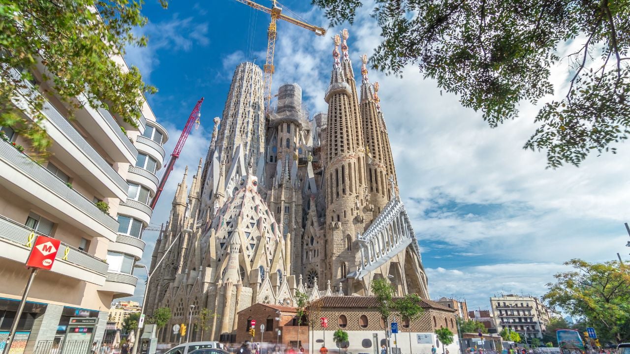 Sagrada Familia Unveiled: Timelapse Hyperlapse of the Iconic Roman Catholic Church in Barcelona, Spain. Autumn's Palette Paints the Scene with Lush Green Trees and a Dynamic Blue Cloudy Sky