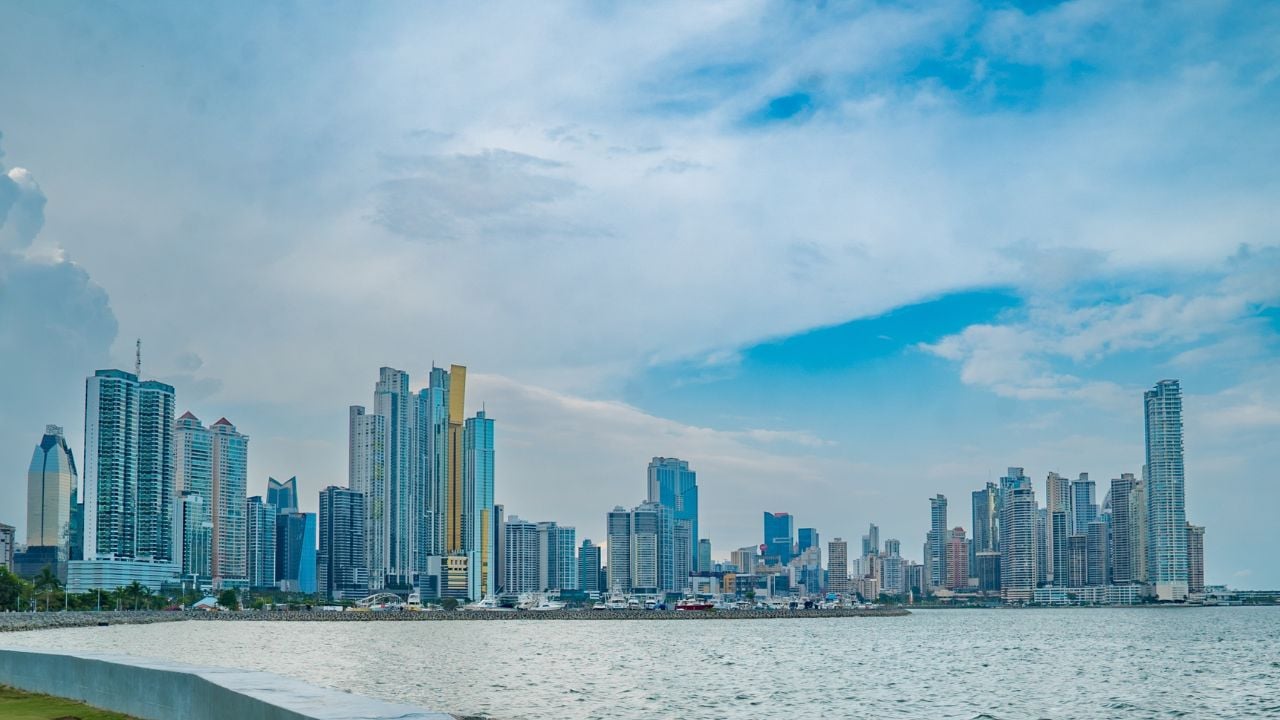 A daytime view of Panama City’s modern skyline seen from a waterfront promenade, with tall skyscrapers reflecting in the calm water and a palm tree adding a tropical touch to the urban landscape.