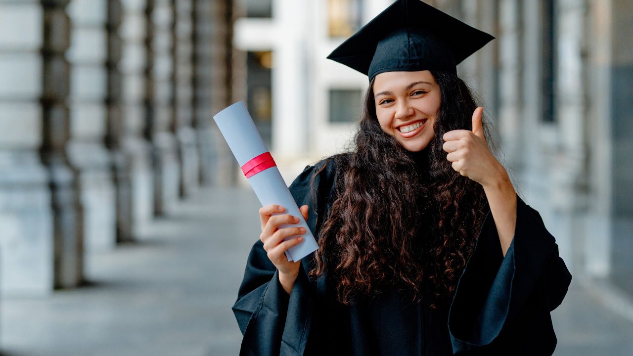 Young latin woman wearing graduation gown holding diploma and showing thumb up, celebrating successful graduation