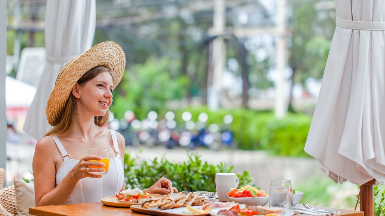Young smiling woman on breakfast in outdoor restaurant in hotel. Female traveler in straw hat drinking orange juice in modern cafe. Summer vacation