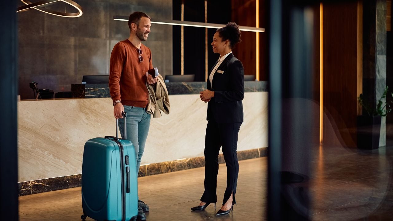 Happy man with a suitcase communicating with African American female concierge in hotel hallway.