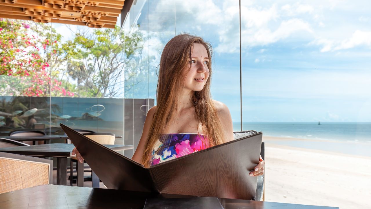 Dining by the sea, young adult engages with menu at beachside eatery, wooden interior with ocean backdrop.