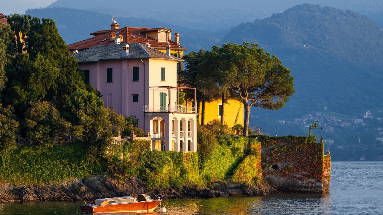 Lakeside scene featuring vibrant villas nestled on shore at Varenna, lake Como, Italy, with small boat floating on the water. Golden sunset light casts warm glow on buildings and landscape