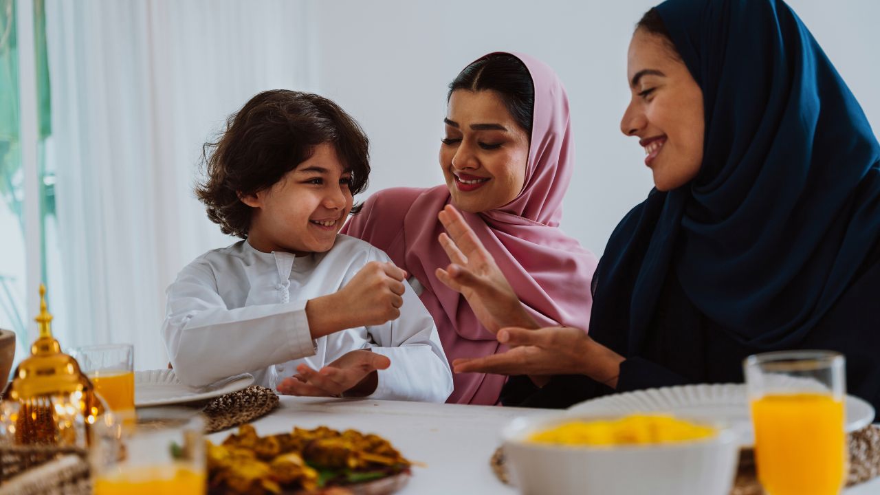 Emirati family at home. Muslim family — women in hijabs and a young boy laugh and play a game while sharing a festive meal at home, joyful eid moment