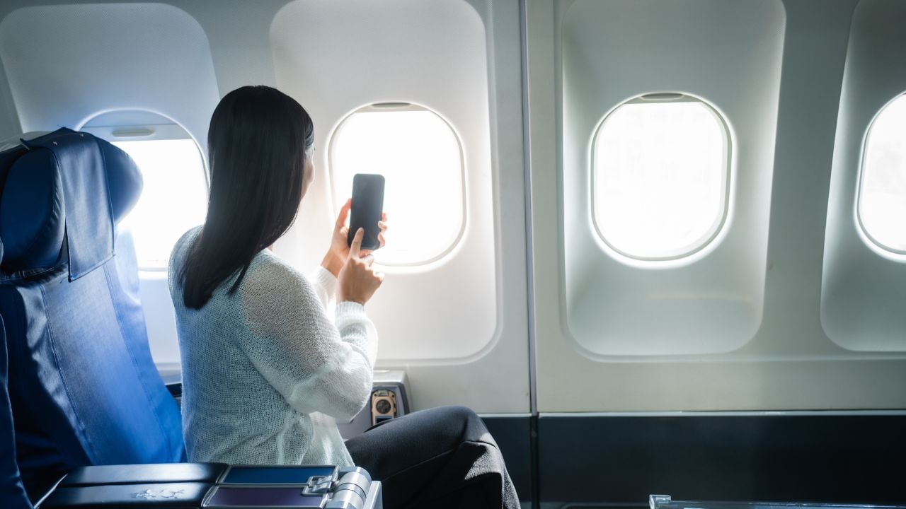 A young Asian female airplane passenger sits by the window during the flight, holding her boarding pass, with her carry-on luggage stored overhead, wearing her seatbelt, and enjoying the journey.