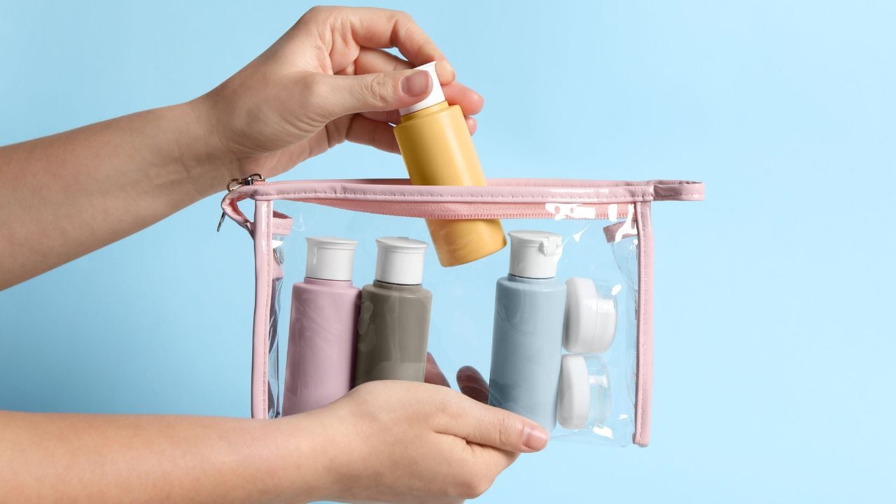 Cosmetic travel kit. Woman putting small bottle with personal care product into plastic bag against light blue background, closeup