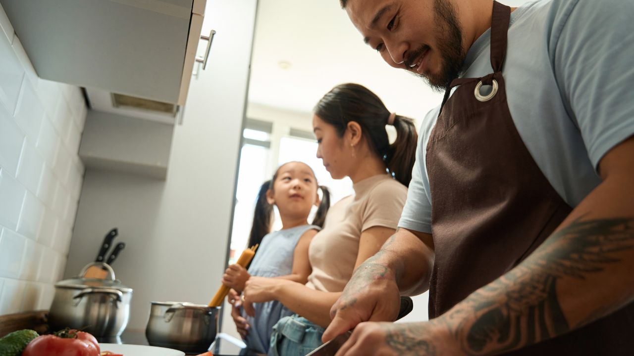 Man cuts tomatoes while woman and a girl cook spaghetti