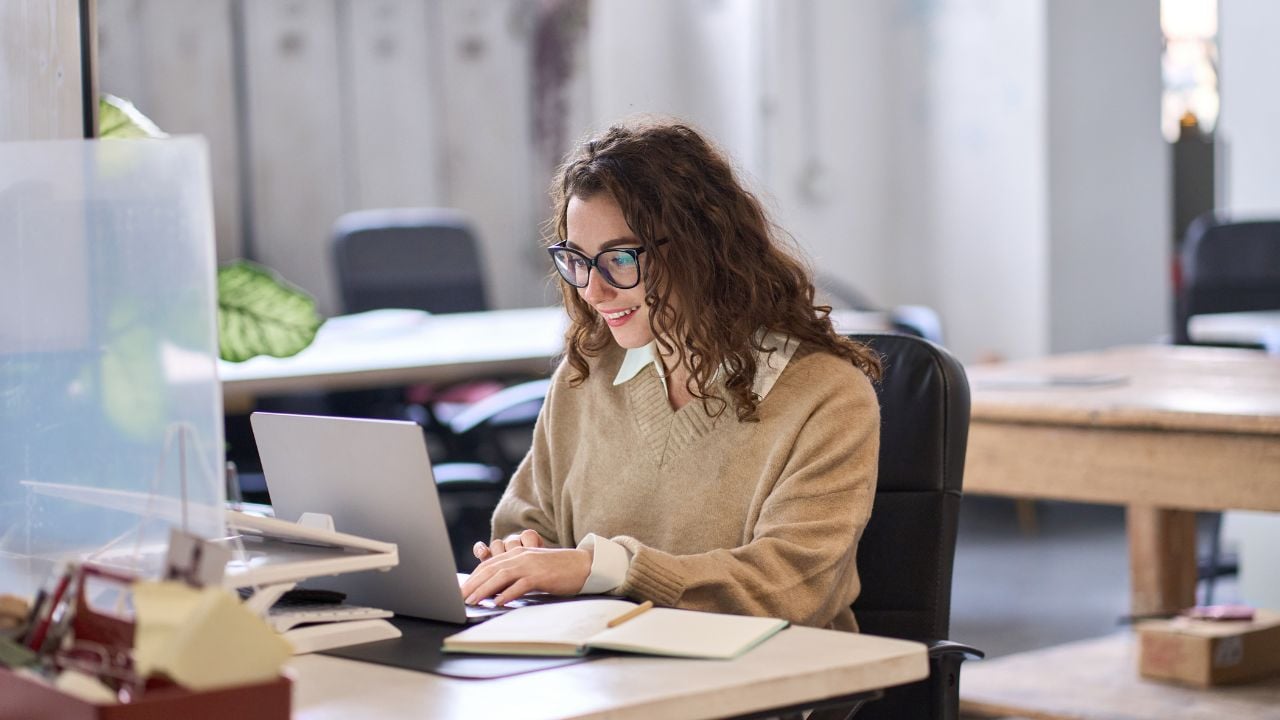 Young happy professional business woman employee sitting at desk working on laptop in modern corporate office interior. Smiling female worker using computer technology typing browsing web.