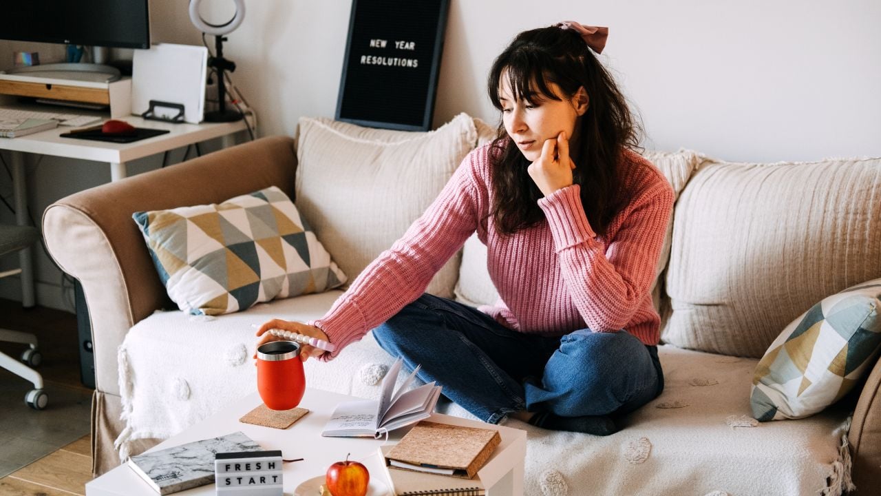 Young adult woman sits cross-legged on sofa holding red coffee mug contemplating personal goals. New Year resolutions, self reflection, mental health, goal setting.