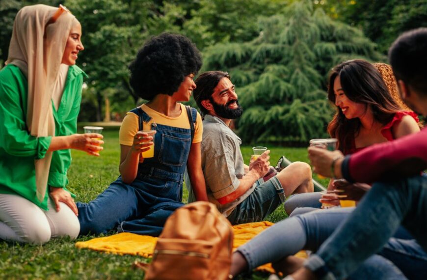 A group of multicultural friends are gathered in the park sitting on the blanket on the lawn enjoying each other's company and having fun.