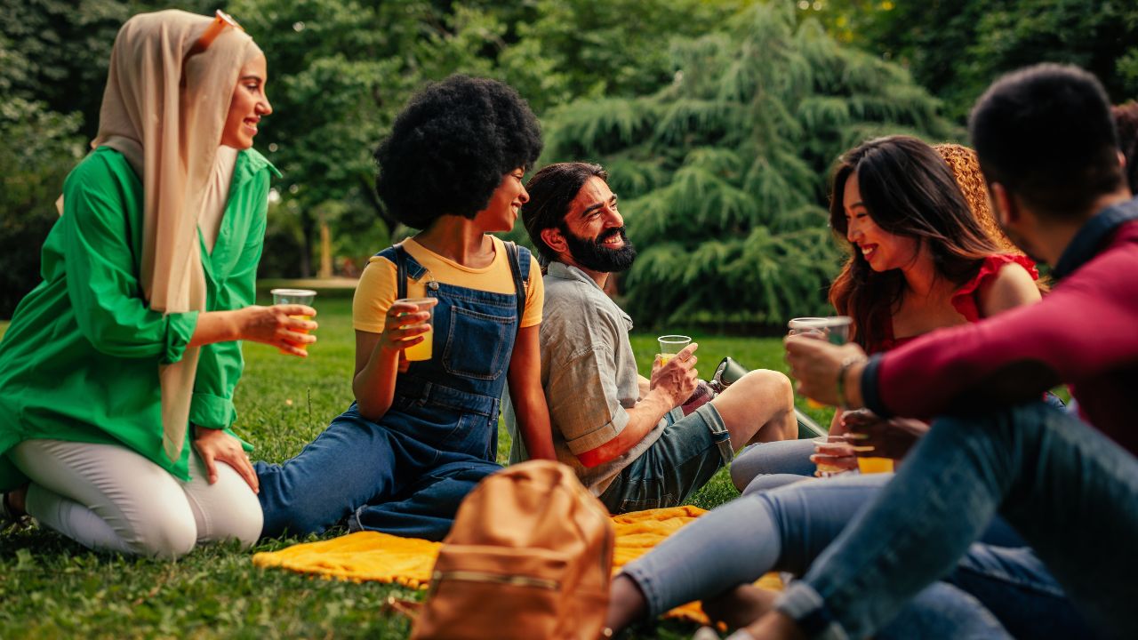 A group of multicultural friends are gathered in the park sitting on the blanket on the lawn enjoying each other's company and having fun.