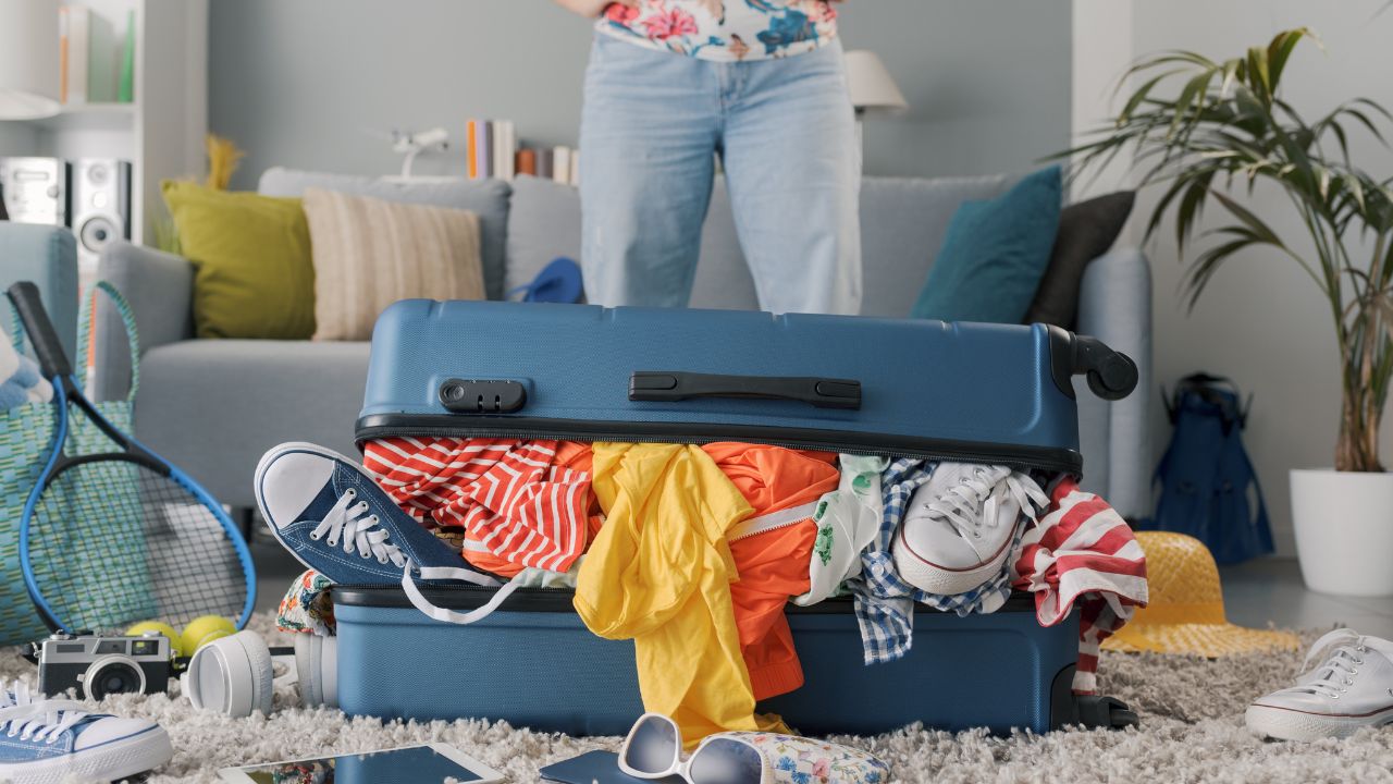 Disappointed woman packing for a journey: she is standing in front of her suitcase overfilled with clothes, travel and vacations concept
