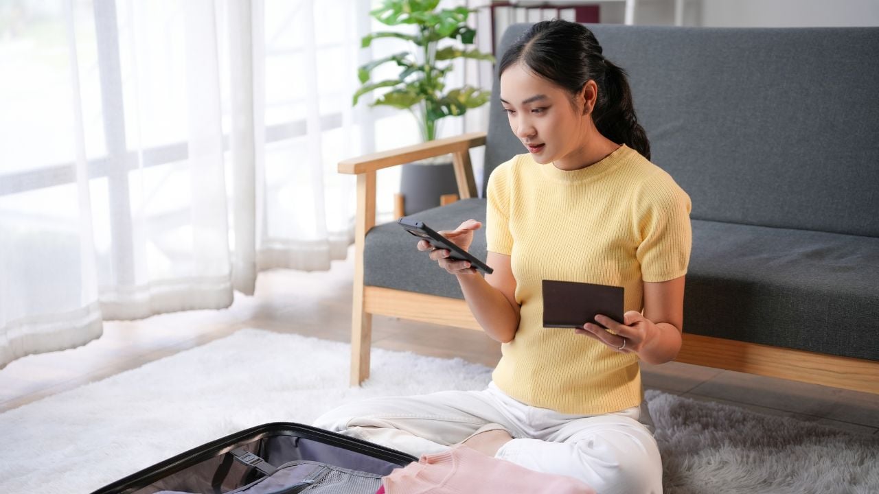 Young woman is packing her luggage while checking her passport and using a smartphone, possibly booking a taxi or browsing travel tips