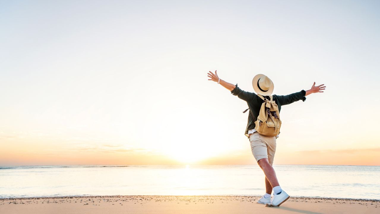 Happy man wearing hat and backpack raising arms up on the beach at sunset - Delightful man enjoying peaceful moment walking outdoors - Wellness, healthcare, traveling and mental health concept