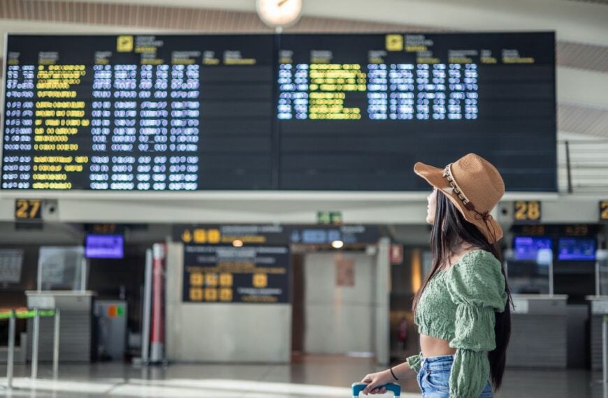 Hispanic girl looking at flight screens in airport terminal while carrying her luggage copy space