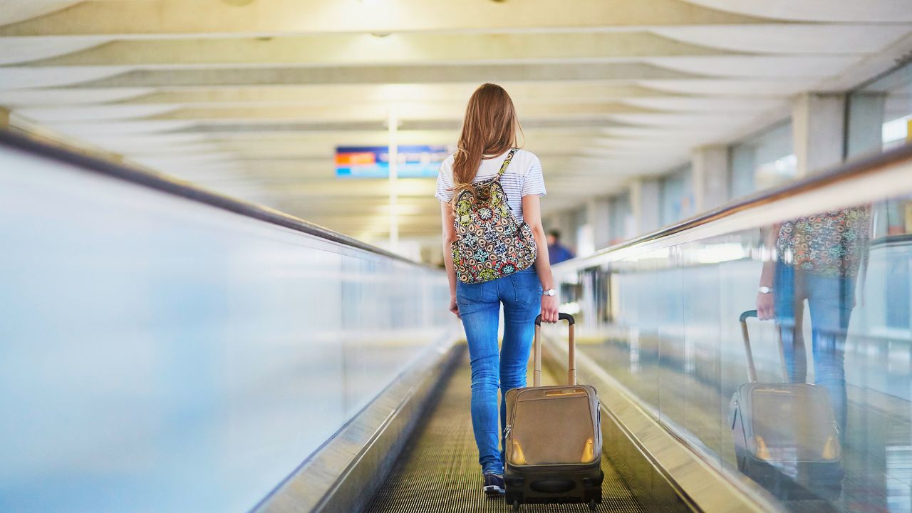 Beautiful young tourist girl with backpack and carry on luggage in international airport, on travelator
