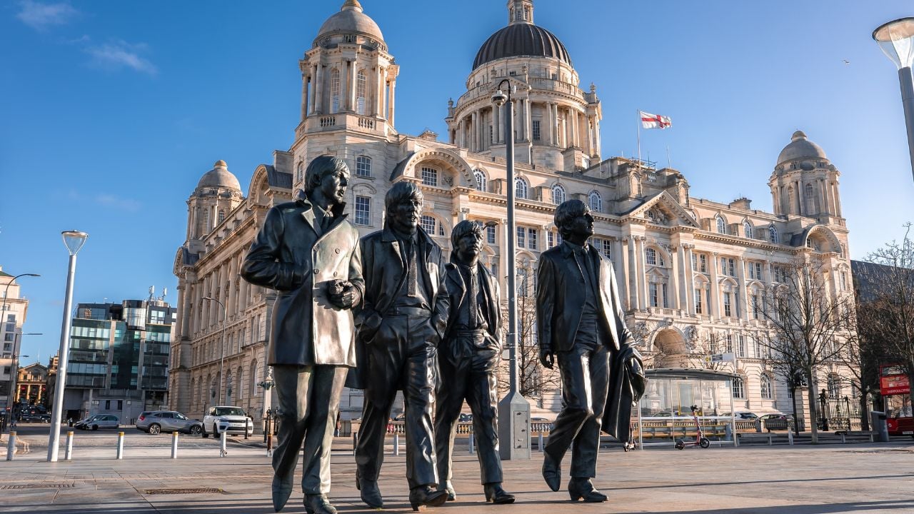 Liverpool, UK. May 1, 2025. Low angle shows bronze Beatles statues walking on Pier Head, Liverpool. Port of Liverpool Building and Union flag stand behind. Winter light casts long shadows.