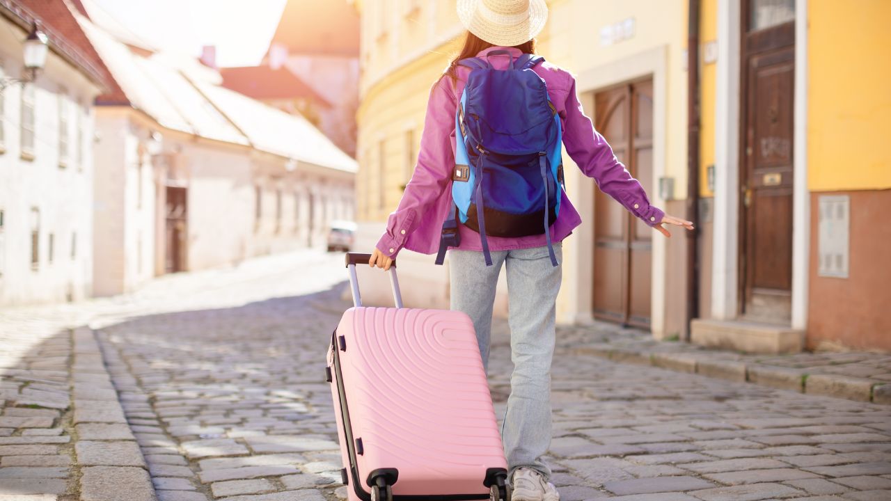 Woman traveler with pink suitcase wandering on a sunny cobblestone street in a quaint European town, embodying adventure and discovery, back view