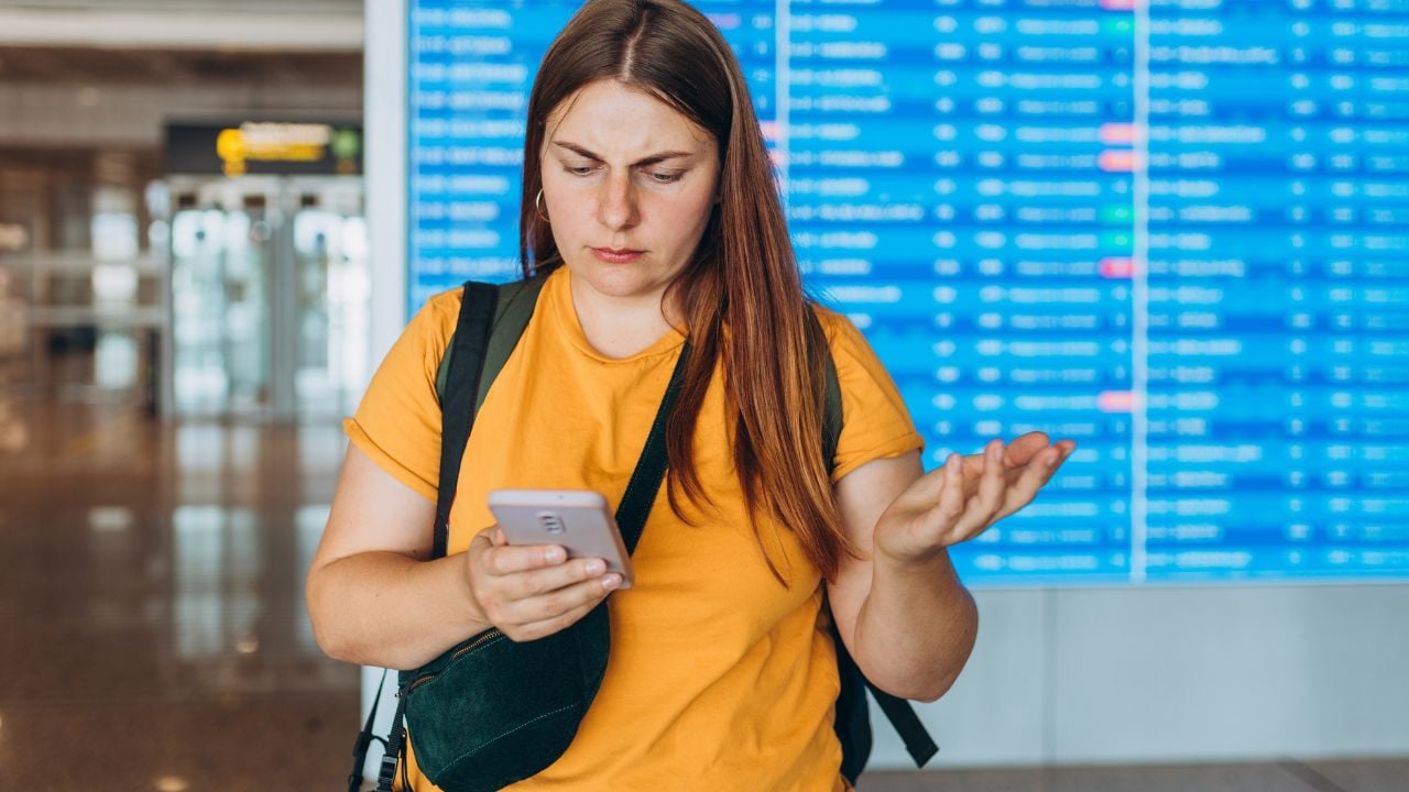Perplexed angry 30s woman holding cellphone in hands, checking Trip Destination on Internet near information board. Delayed flight. Receives Shockingly Bad News, Misses Flight.