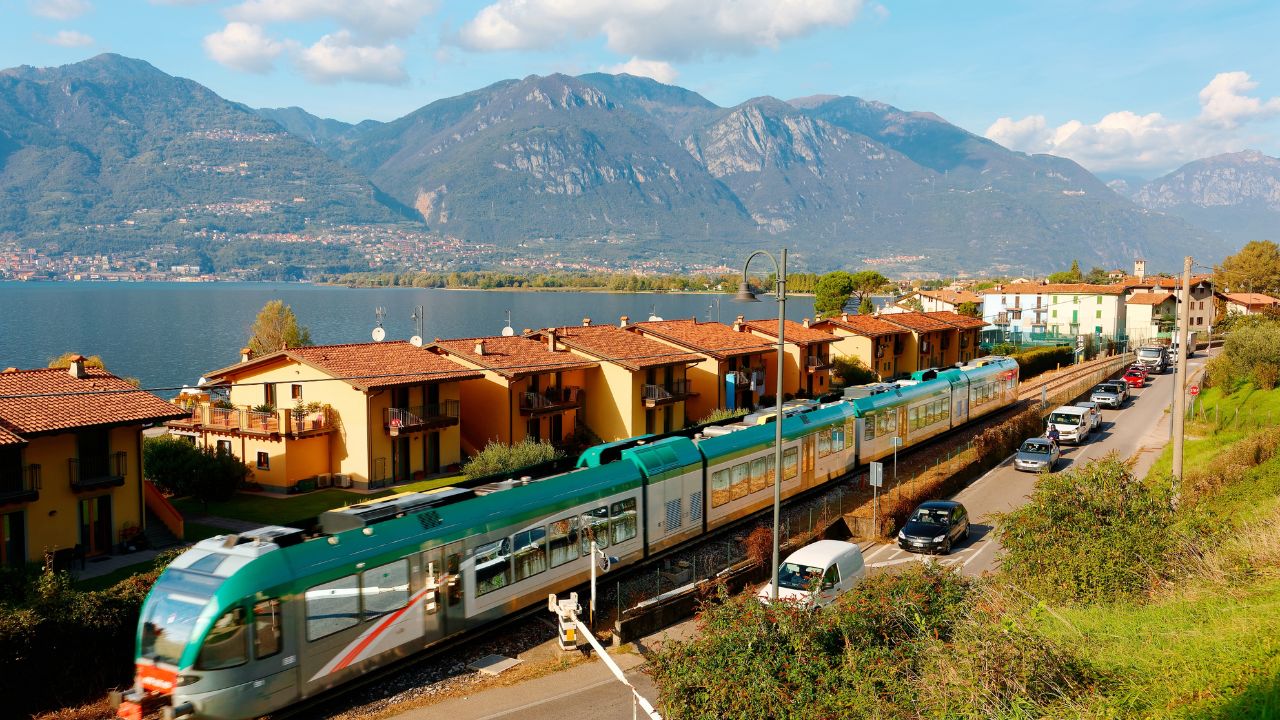 Scenery of beautiful Lake Iseo under blue summer sky, with a regional train traveling by a row of lakeside houses and cars stopping before the railroad crossing, in Pisogne, Lombardy, northern Italy