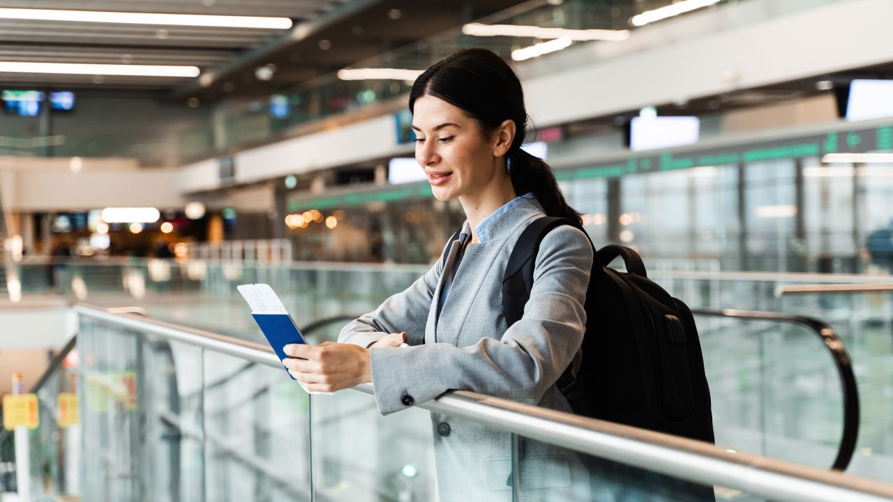 Woman holding passport, boarding pass and smartphone standing near window at airport lobby and waiting for departure. Tourist in airport is standing near panoramic window waiting for security check