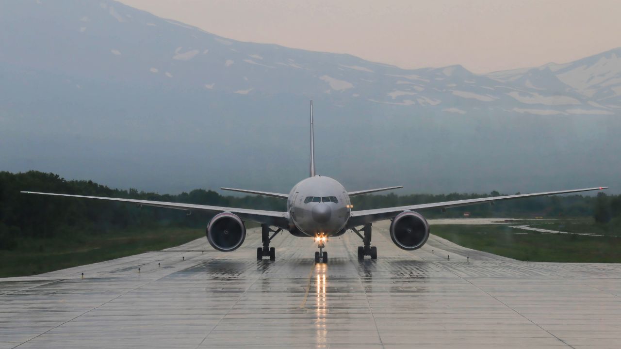 Frontal view of Aircraft coming in for Landing against background of Snow-capped Mountain Peaks on Rainy cloudy day. The Light Path from Headlights creeps along the Wet runway towards the airport.