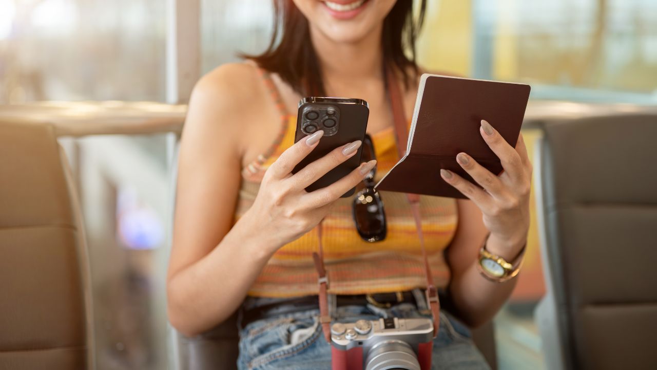 A cropped image of a beautiful young Asian female tourist is holding her smartphone and passport, checking her flight information on her phone while sitting at a waiting seat in the airport terminal.