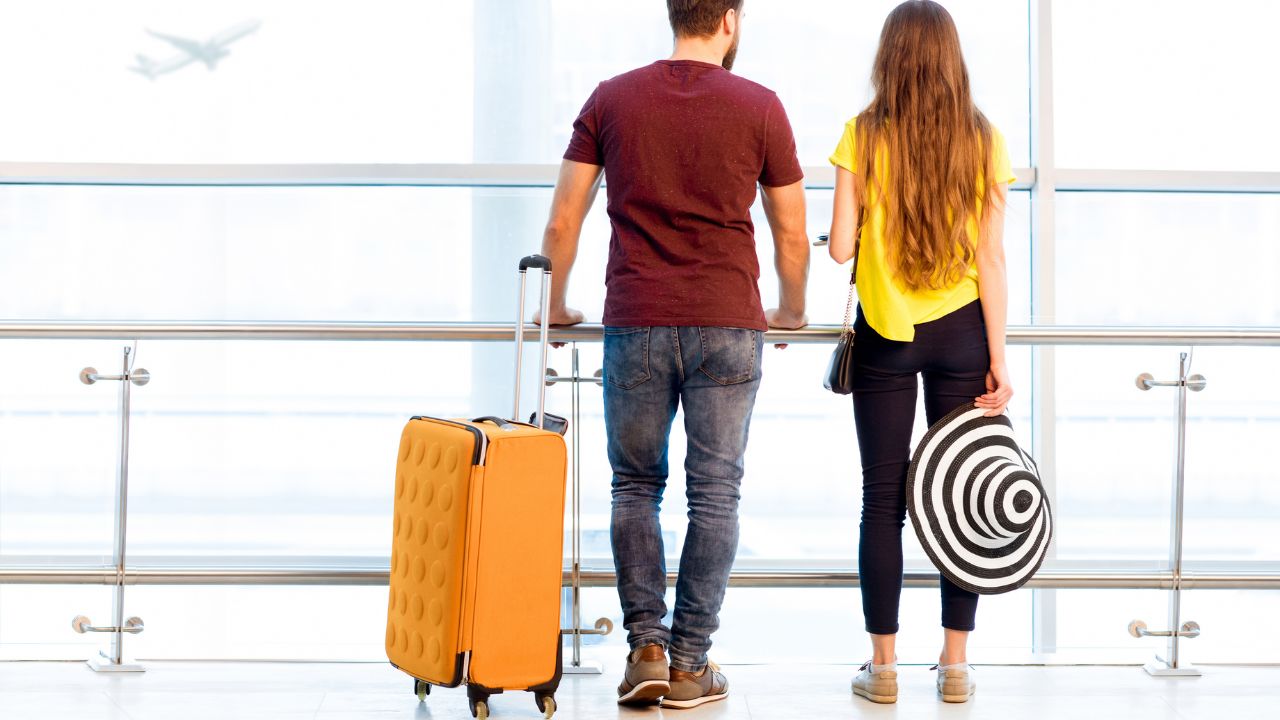 Young couple in colorful t-shirts waiting with baggage near the window at the departure area of the airport during their summer vacation