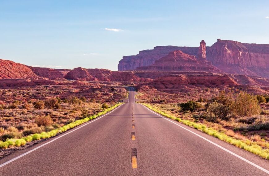 A long, empty road stretches through the red desert towards the stunning rock formations of Monument Valley. The scene captures the vastness and beauty of the Arizona landscape.