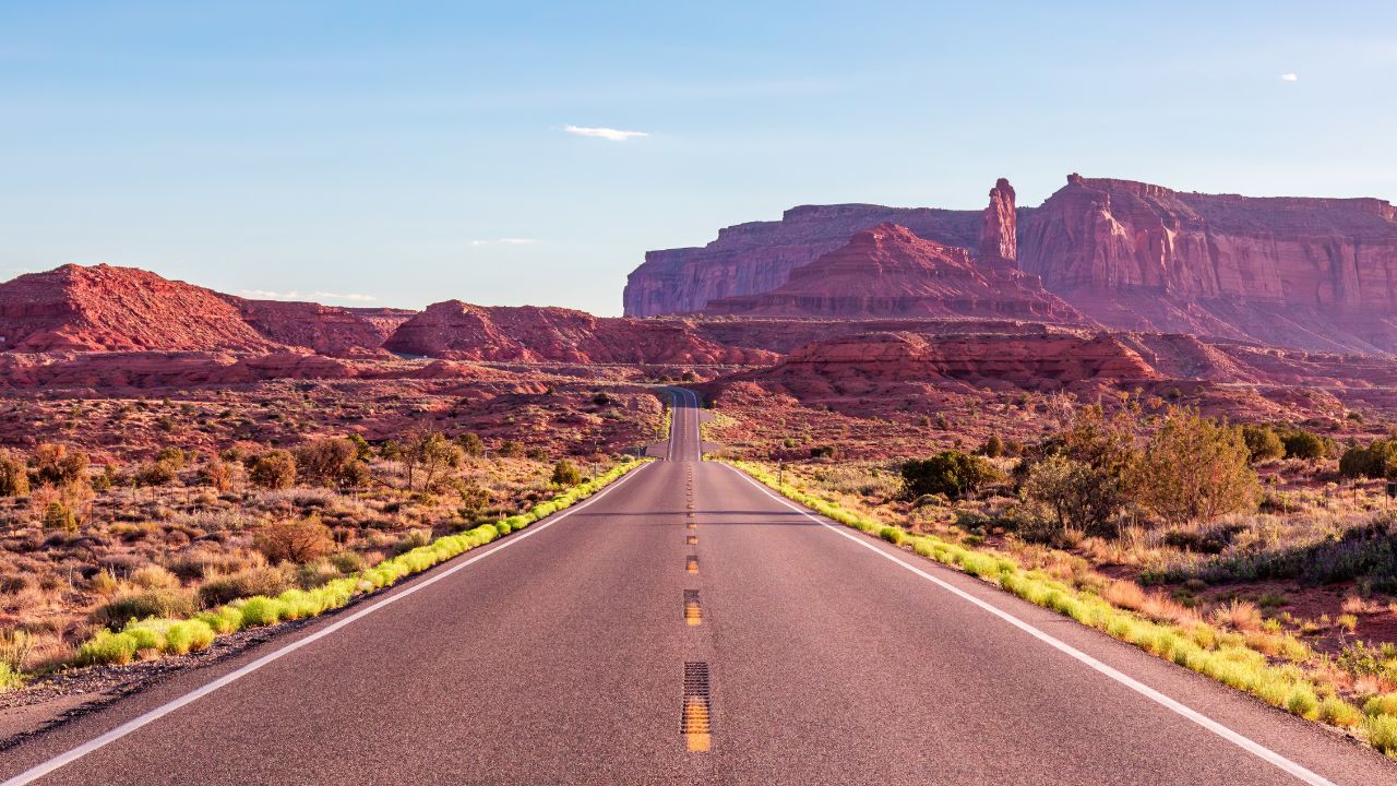 A long, empty road stretches through the red desert towards the stunning rock formations of Monument Valley. The scene captures the vastness and beauty of the Arizona landscape.