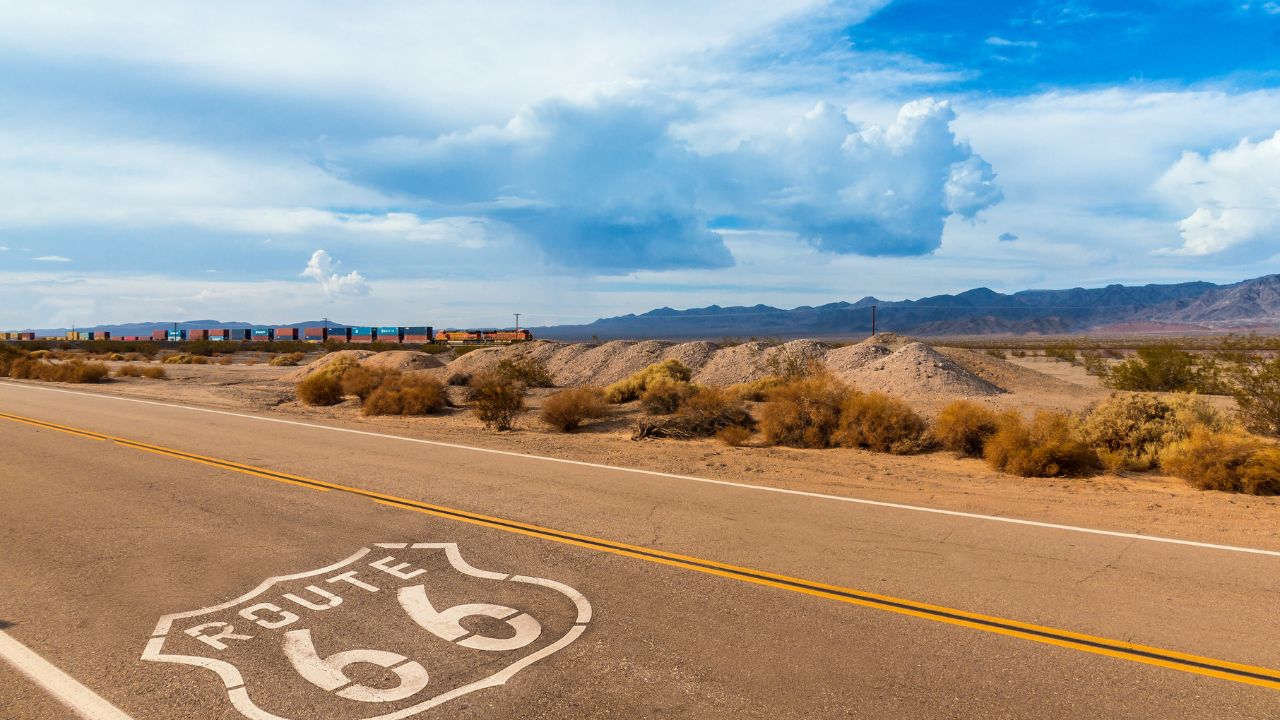 U.S. Route 66 highway, with sign on asphalt and a long train in the background, near amboy, california. Located in the mojave dessert