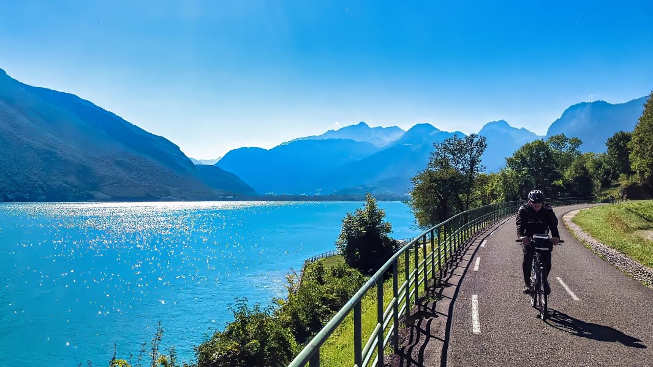 Annecy, France - July 04, 2016 : A cyclist enjoys a scenic ride beside a clear blue lake in Haute Savoie surrounded by majestic Alps on a sunny day.