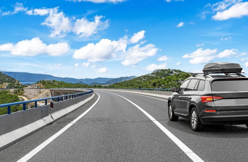 A modern SUV with a roof box drives along a highway through a scenic mountain landscape under a bright blue sky with clouds.