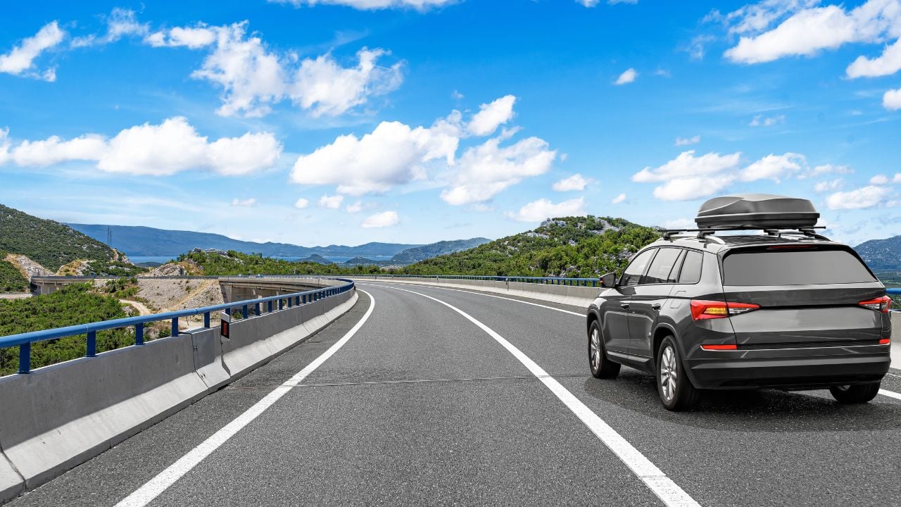 A modern SUV with a roof box drives along a highway through a scenic mountain landscape under a bright blue sky with clouds.