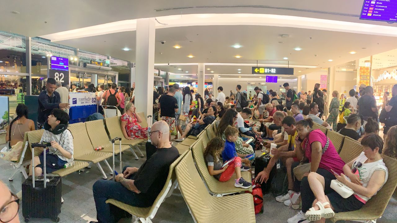 A crowded airport terminal with people sitting on chairs and waiting for their flights. Scene is busy and hectic, with people rushing to catch their flights and others waiting patiently