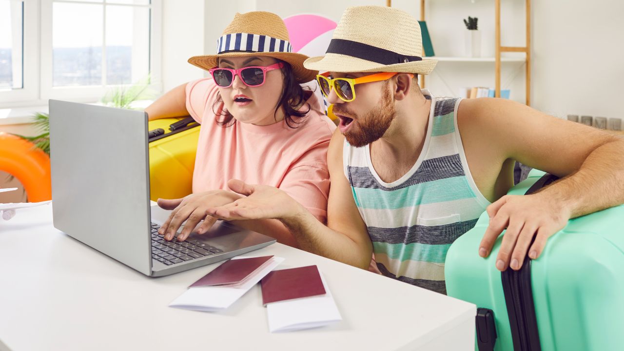 Funny tourists couple booking hotel online using laptop. Shocked husband and overweight wife in summer clothing and hats sitting on sofa at home with packed suitcases planning vacation trip