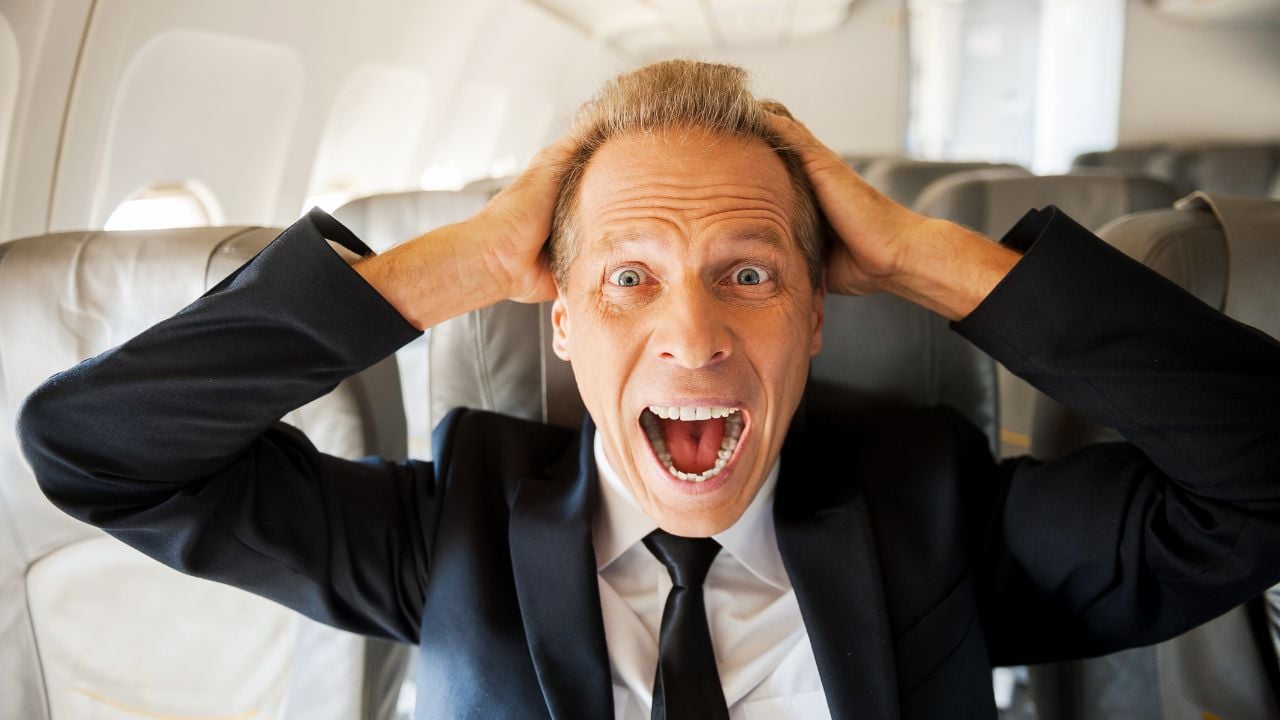 Fear of flight. Shocked mature businessman touching his head with hands and looking at camera while sitting at his seat in airplane