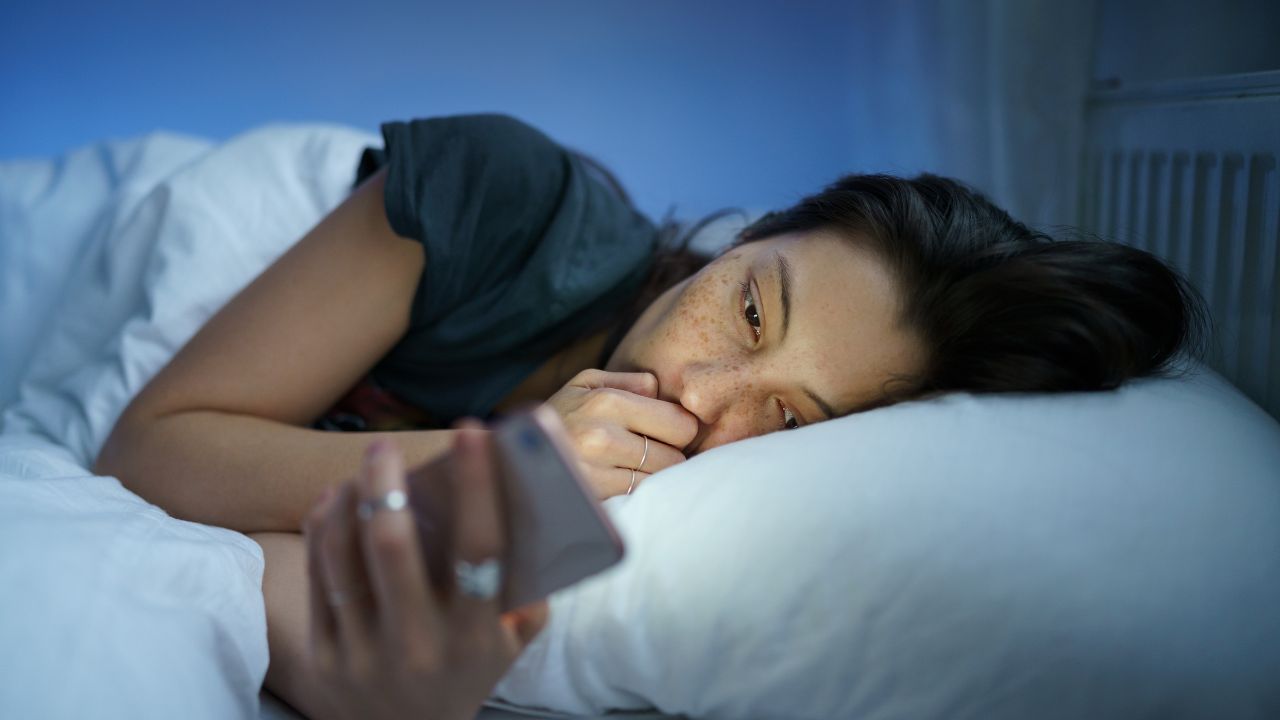 Depressed sleepless young woman follow cheating ex-boyfriend in social media lying tired in bed and suffering from insomnia. Anxious female with sleep disorder look at smartphone screen in dark room