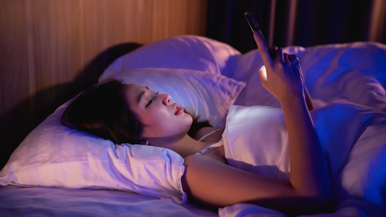 Asian woman using mobile phone smartphone laying on the bed in the bedroom. Sleepy exhausted, can not sleep. Insomnia, addiction concept. Women scrolling social networks on mobile dark bedroom.