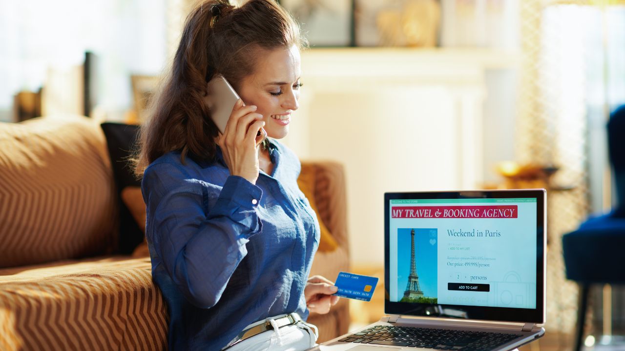 happy young woman with opened on laptop online travel & booking agency site holding credit card talking on a smartphone in the modern house in sunny summer day.