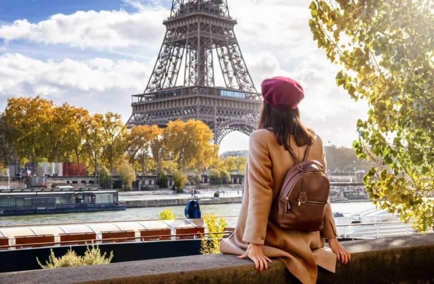 A tourist woman with a beret hat sits at the riverbank of the Seine and enjoys the beautiful view of the Eiffel Tower in Paris, France