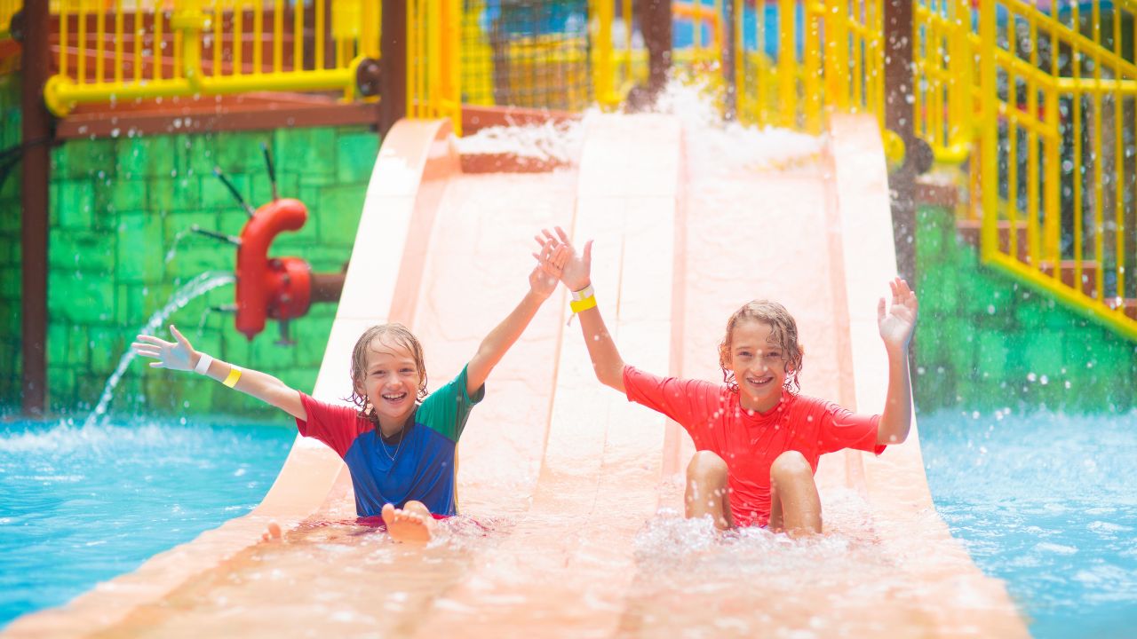 Kids on water slide in outdoor swimming pool. Family in aqua theme park. Children have fun in splash playground. Summer vacation with child. Kid on inflatable tube sliding into a pool.