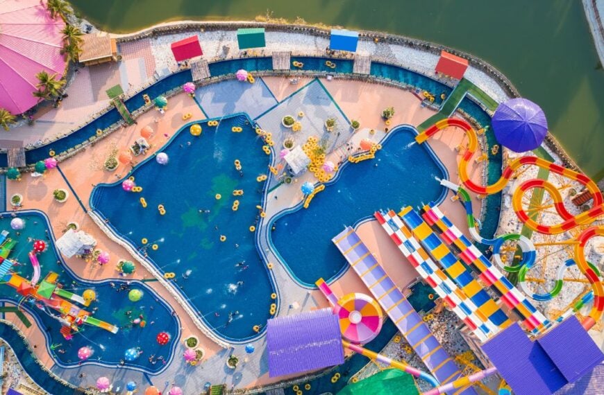 A colorful aerial view shows a large waterpark with slides, pools, and attractions designed for families enjoying a summer day