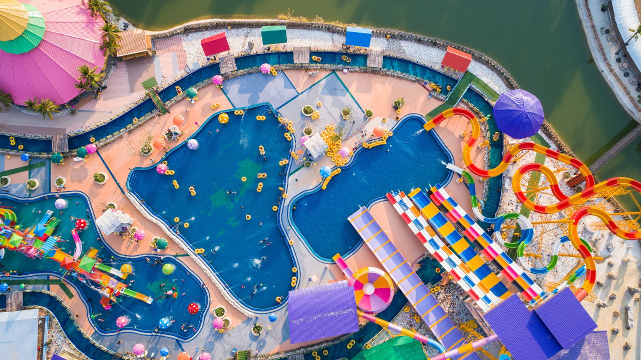 A colorful aerial view shows a large waterpark with slides, pools, and attractions designed for families enjoying a summer day