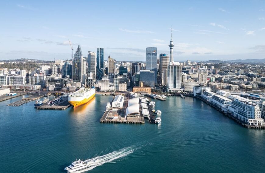 Auckland, New Zealand - August 14 2024: Aerial drone view of a ferry that leaves the Auckland historic ferry terminal with the skyline and a massive car carrier in New Zealand largest city