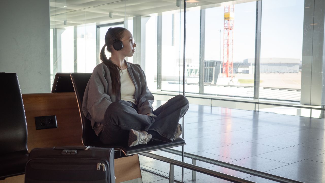 A woman sits cross-legged on an airport bench, listening to music through headphones and looking out the window, suitcase nearby. Concept of peaceful travel, anticipation and solo journey experience.
