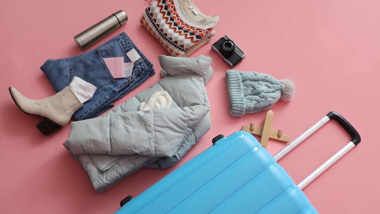Suitcase with warm clothes, thermos and camera on pink background. Winter vacation concept
