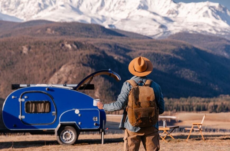Amazing van life travel concept. Young man traveler with hat and coffee enjoying mountain view with blue trailer camper.