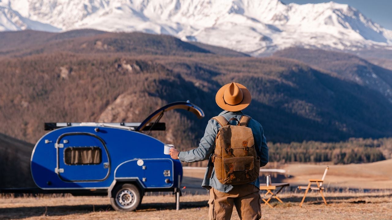 Amazing van life travel concept. Young man traveler with hat and coffee enjoying mountain view with blue trailer camper.